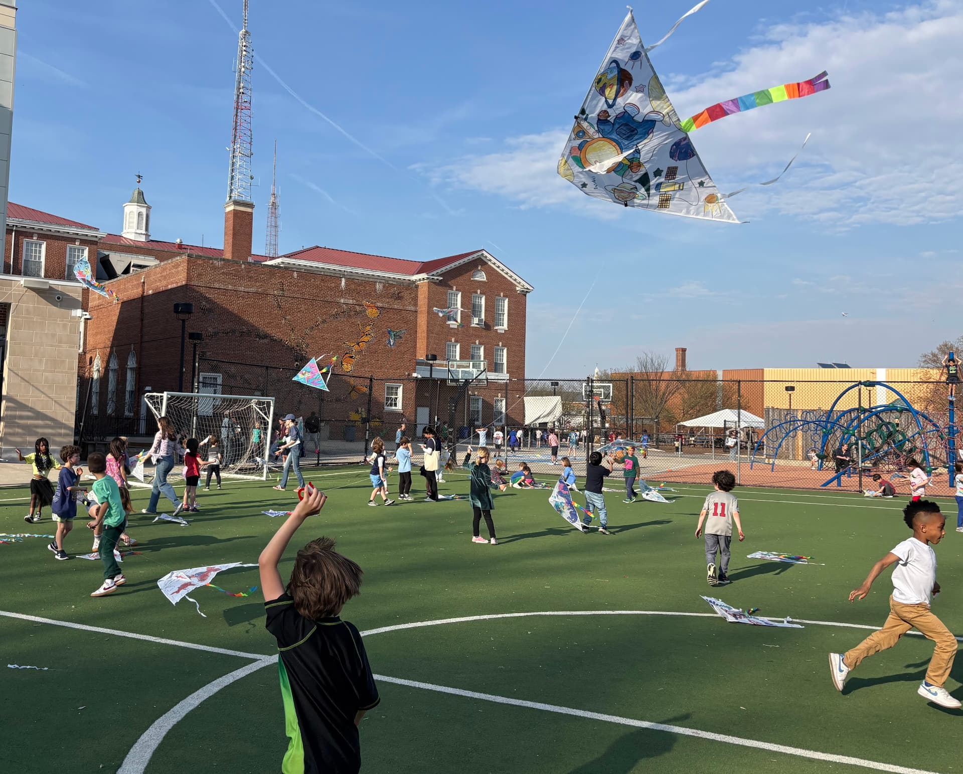 A large group of Janney+ kids flying colorful kites together on the school turf field on a sunny day