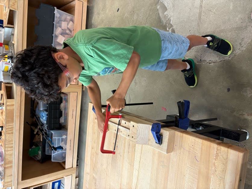 A student in safety glasses sawing a piece of wood at a workbench
